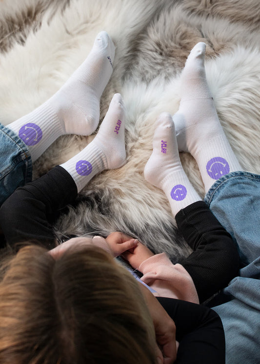 Zwei Paar weiße J.Clay-Socken - ein Erwachsener, ein Baby - jeweils mit einem lila Smiley-Gesicht. Perfekt für Partner-Look oder Mini Me Momente! Auf den Babysocken steht "J.Clay" in lila in der Nähe der Zehen. - Mama Kind Packs