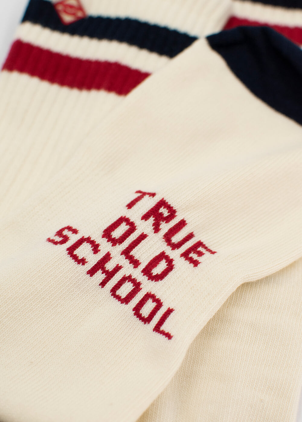 Eine Nahaufnahme der cremefarbenen Tennissocken von J.Clay mit rotem "TRUE OLD SCHOOL"-Schriftzug, die neben gerippten Socken mit klassischen marinefarbenen und roten Streifen zu sehen sind.