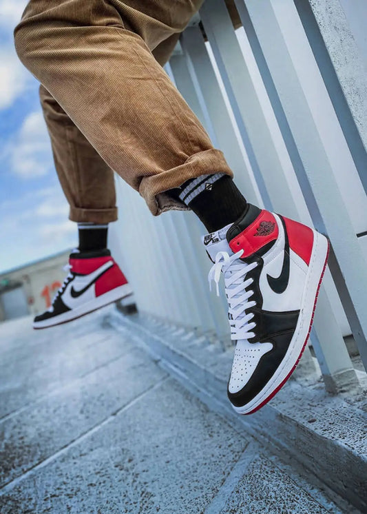 Person in brauner Hose, Basic Black Socken von J.Clay und Nike Turnschuhen steht an einem weißen Zaun.