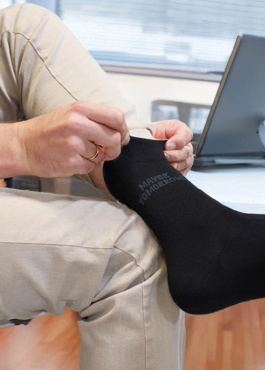 Jemand trägt eine beigefarbene Hose und zieht eine schwarze Business-Socke von J.Clay an, auf der "MAYBE TOMORROW" steht, während er neben einem Laptop auf einem Schreibtisch sitzt.