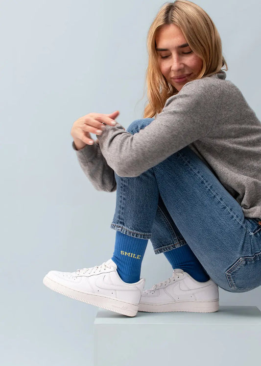 Frau in Jeans und grauem Pullover, trägt J.Clay Smile Tennissocken und weiße Turnschuhe, sitzt entspannt.