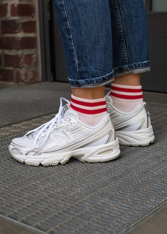 Ein Damen-Outfit mit J.Clay farbenfroh Sneaker Socken mit roten Streifen, gepaart mit weißen Turnschuhen mit silbernen Details und blauen Jeans mit Bündchen, stehend auf einer strukturierten Matte in der Nähe einer Ziegelwand und einer Tür.