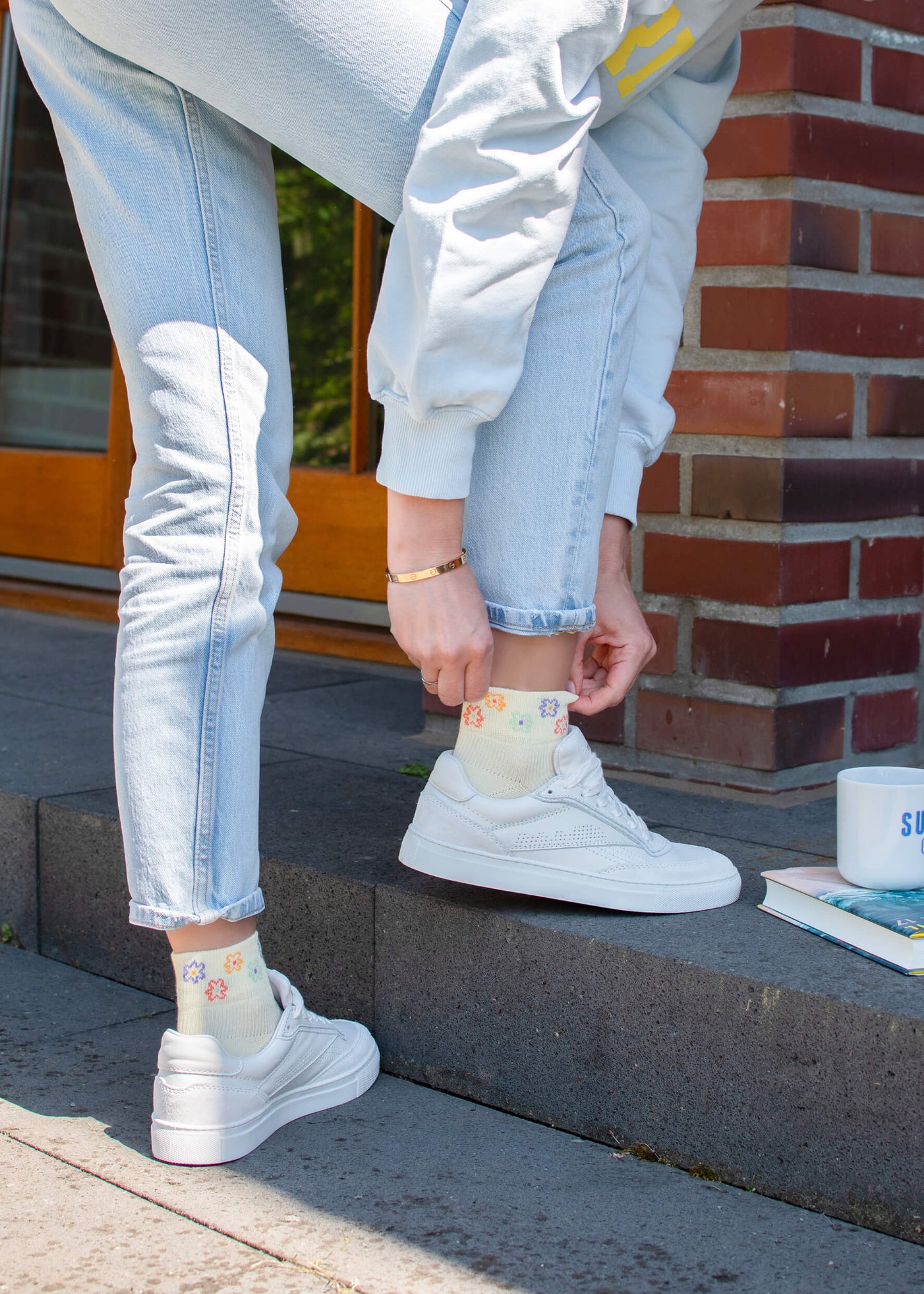 Eine Person in hellen Jeans, einem hellen Sweatshirt und weißen Turnschuhen passt ihre J.Clay Sneaker Socken - bunt gemusterte Socken - an, während sie auf Steinstufen neben Büchern und einem weißen Becher steht.