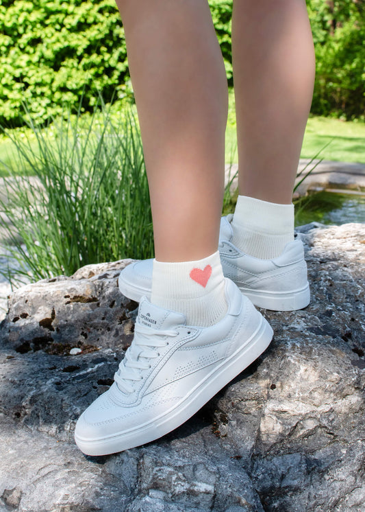 Eine Person steht im Freien auf einem Felsen und trägt J.Clay Sneaker Socken - weiße Socken mit einem kleinen roten Herz - gepaart mit weißen Turnschuhen, umgeben von grünem Gras und Pflanzen im Hintergrund.
