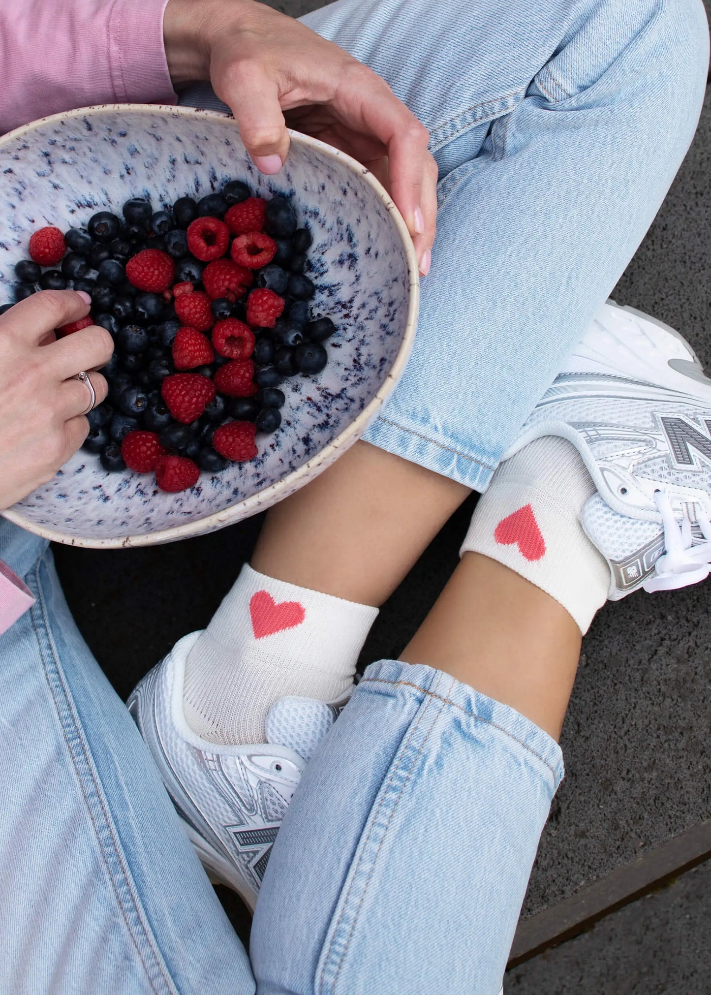 Eine Person in hellblauen Jeans und weißen Turnschuhen sitzt im Schneidersitz und trägt J.Clay atmungsaktive Sneaker Socken mit Herz-Motiv am Knöchel. Ein rosafarbener Ärmel ist sichtbar, während sie eine Schale mit Blaubeeren und Himbeeren hält.