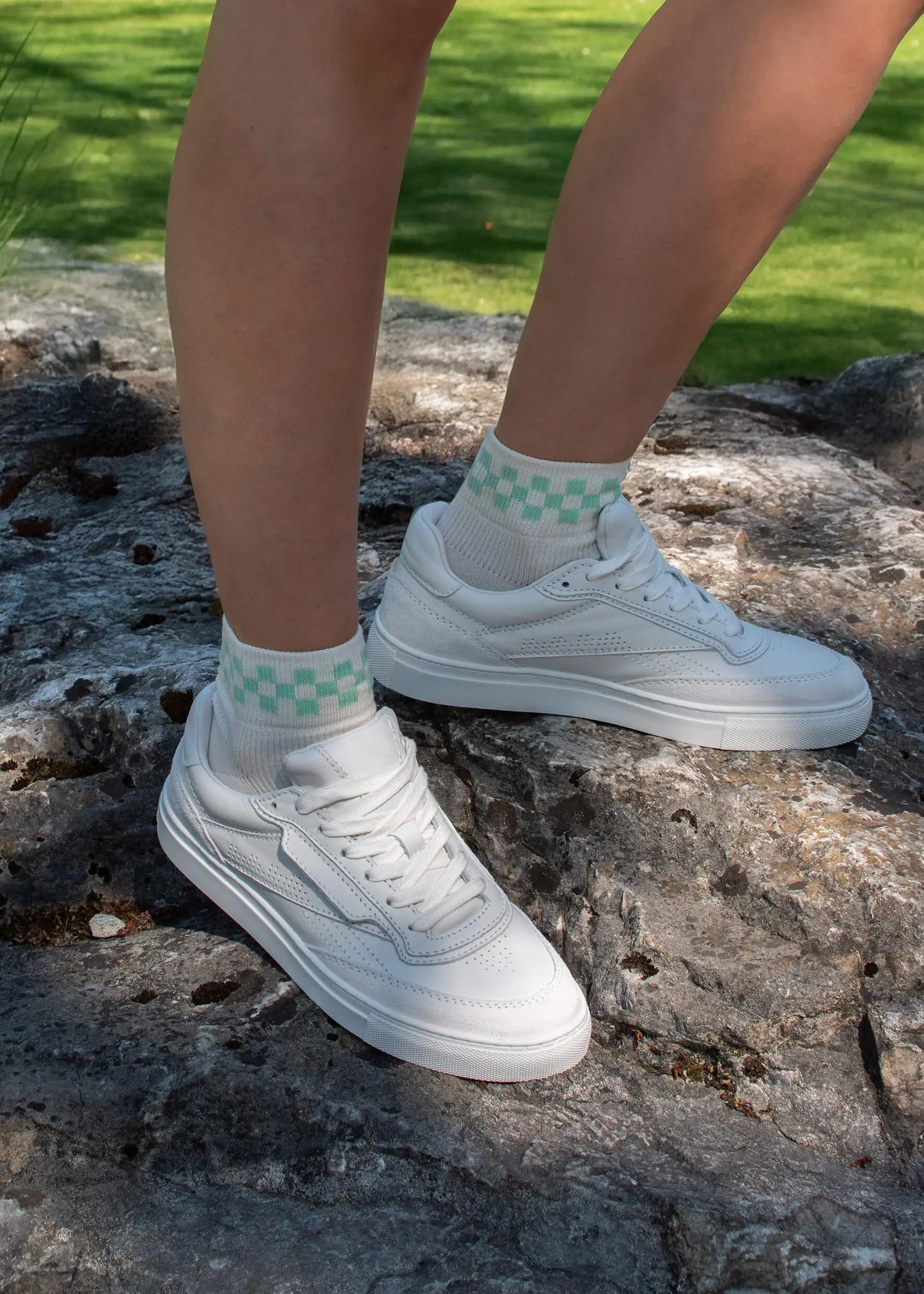 Eine Person trägt J.Clay weiße Sneaker Socken aus hochwertiger Baumwolle mit grünem Karomuster und steht auf einem großen grauen Felsen im Freien, während Gras im Hintergrund zu sehen ist.