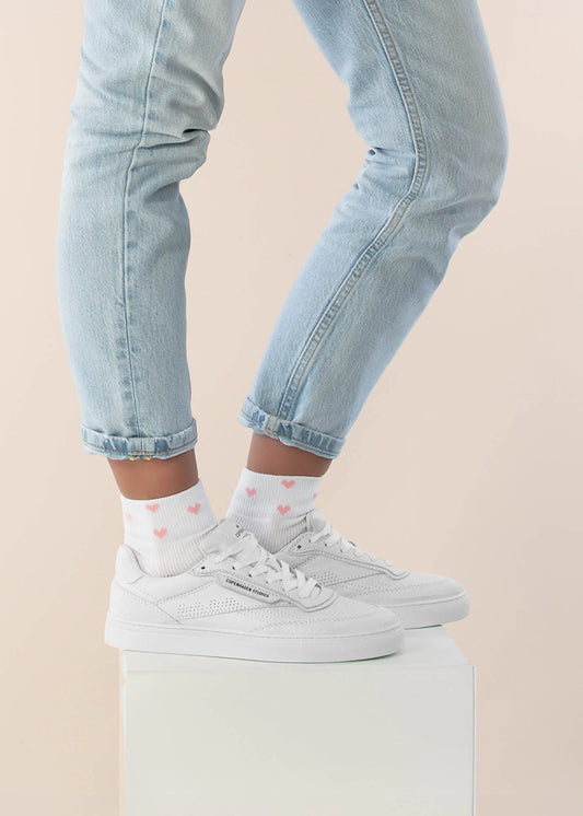 Eine Person in hellblauen, hochgekrempelten Jeans, weißen Turnschuhen und J.Clay Sneaker Socken mit rosa Herzmustern steht auf einem weißen Podest vor einem hellen Hintergrund.