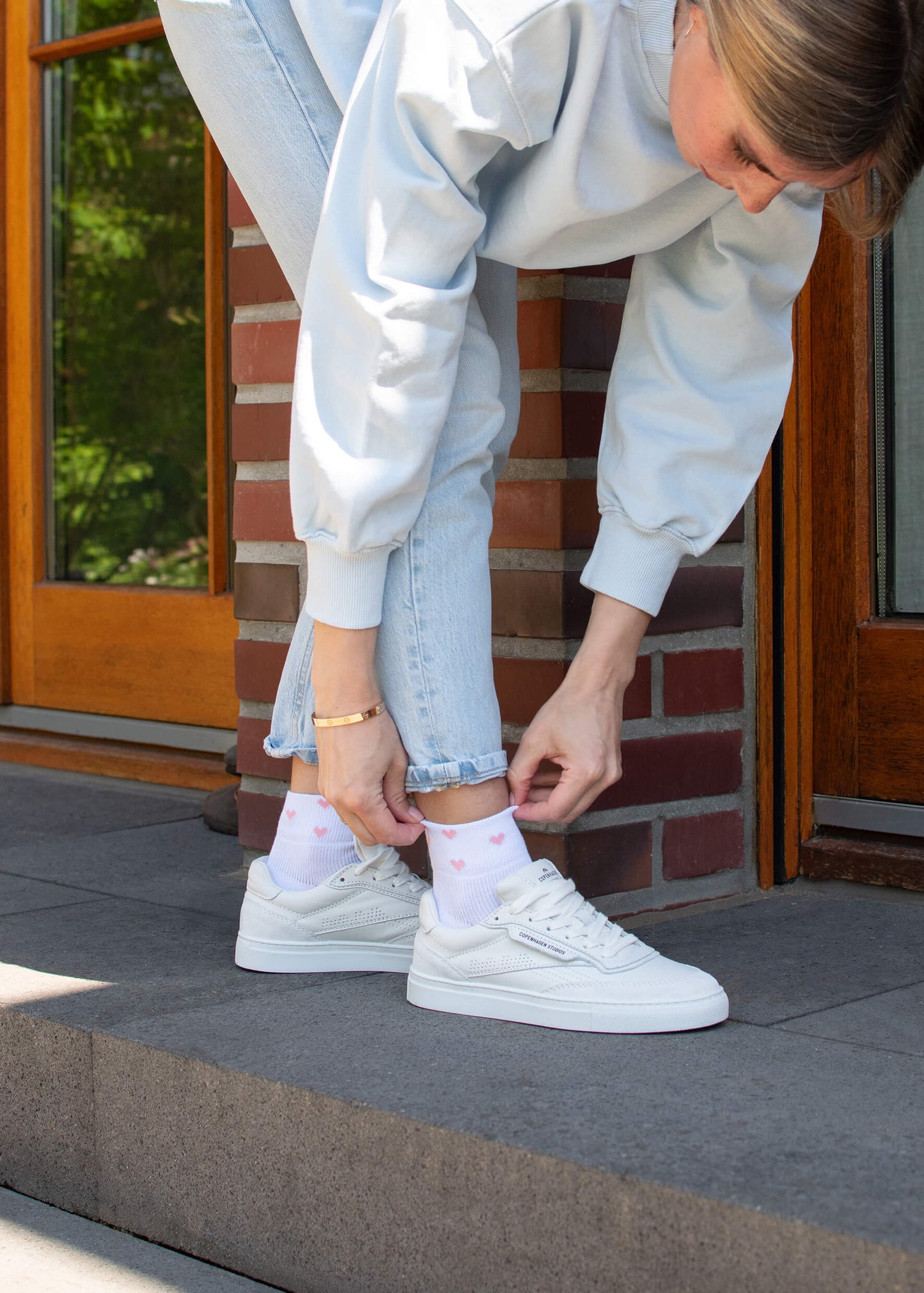 Eine Person in hellblauen Jeans und weißem Sweatshirt passt ihre atmungsaktiven J.Clay weiße Sneaker Socken mit pinken Herzen an, während sie auf einer grauen Türschwelle vor einer Ziegelwand und einer Holztür steht.