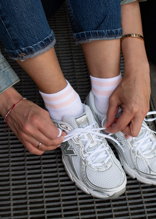 Eine Person in Stulpenjeans, J.Clay Sneaker Socken mit rosafarbenen Streifen und weißen Turnschuhen aus atmungsaktivem Material bindet ihre Schnürsenkel, während sie auf einem Metallgitter sitzt, und trägt Armbänder an beiden Handgelenken und einen Ring an einem Finger.