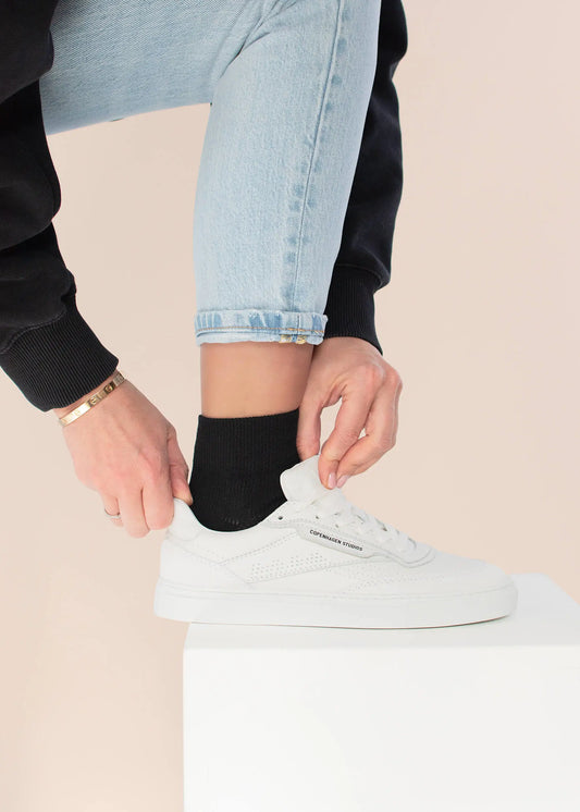 Eine Person in hellblauen Jeans und einem schwarzen Pullover passt die Schnürsenkel von weißen J.Clay Sneakers an, während sie auf einer weißen Fläche steht. Der Look wird mit schwarzen Socken und einem goldenen Armband abgerundet.