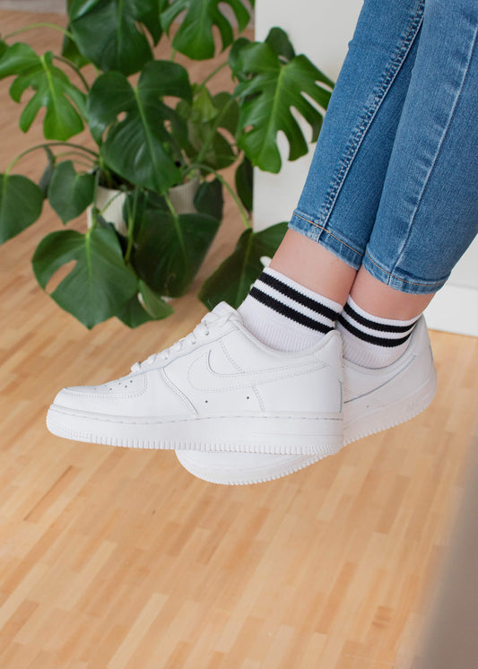 Jemand, der J.Clay Sneaker Socken, weiße Turnschuhe und blaue Jeans trägt, sitzt oder schwebt über einem hellen Holzboden, mit einer grünen Blattpflanze im Hintergrund.