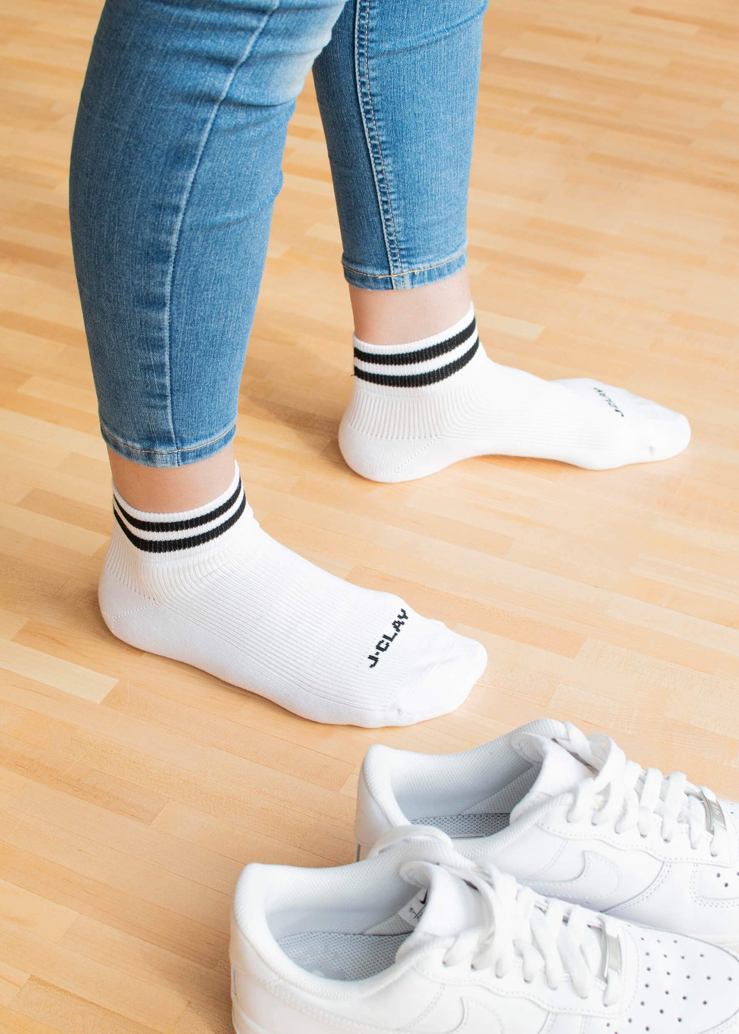 Eine Person, die J.Clay Sneaker Socken - weiße Socken mit schwarzen Streifen - und blaue Jeans trägt, steht auf einem hellen Holzboden neben einem Paar weißer Turnschuhe.