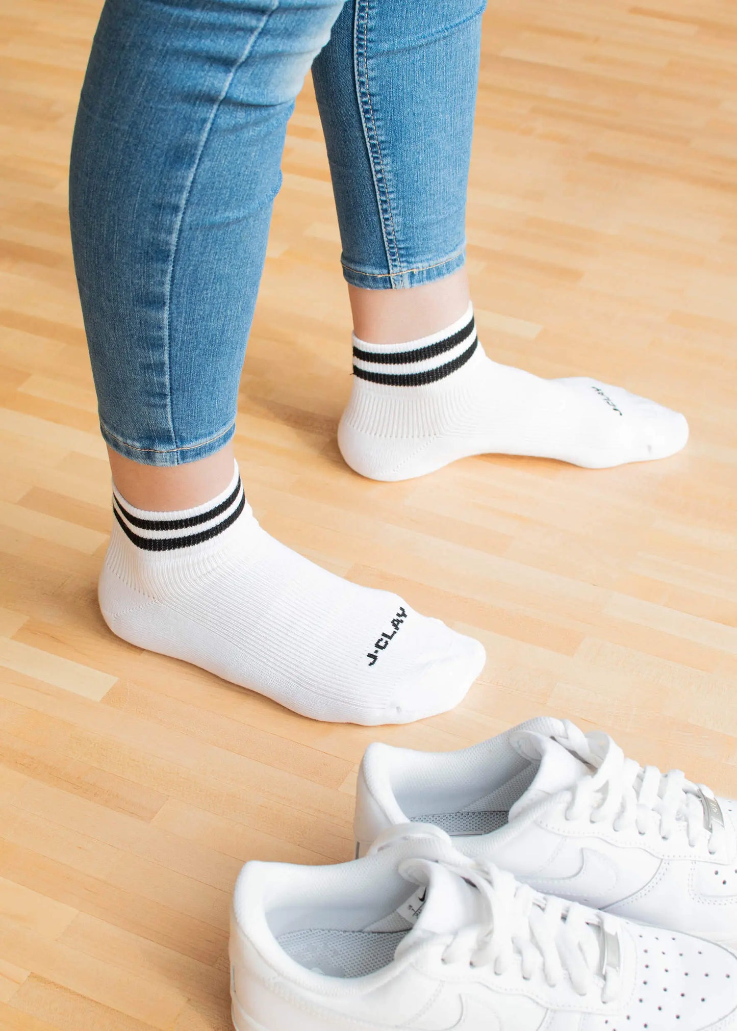 Eine Person, die J.Clay Sneaker Socken - weiße Socken mit schwarzen Streifen - und blaue Jeans trägt, steht auf einem hellen Holzboden neben einem Paar weißer Turnschuhe.