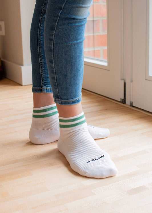 Eine Person, die J.Clay Sneaker Socken - weiß mit grünen Streifen - und blaue Jeans trägt, steht auf einem hellen Holzboden neben einer Glastür. Eine Socke trägt das J.Clay-Logo.