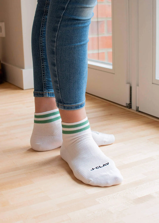 Eine Person, die J.Clay Sneaker Socken - weiß mit grünen Streifen - und blaue Jeans trägt, steht auf einem hellen Holzboden neben einer Glastür. Eine Socke trägt das J.Clay-Logo.
