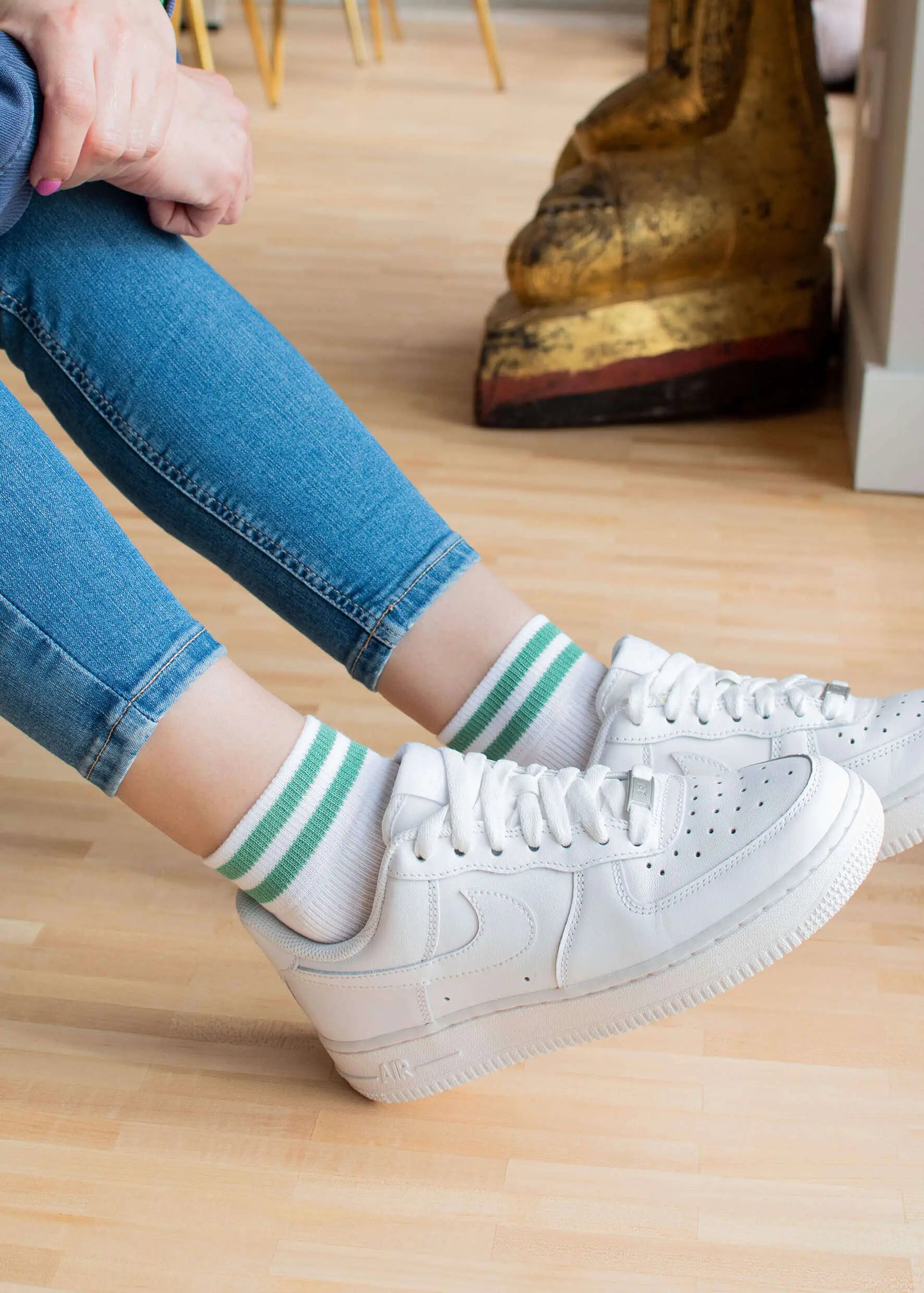 Eine Person in hochgekrempelten Bluejeans und weißen J.Clay-Sneaker-Socken mit grünen Streifen, gepaart mit weißen Turnschuhen, sitzt in einem Innenraum auf einem hellen Holzboden; im Hintergrund ist ein großes Dekorationsobjekt zu sehen.