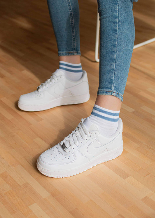 Person in weißen J.Clay Sneaker Socken, weißen Turnschuhen und blauen Skinny Jeans, die auf einem hellen Holzboden steht.