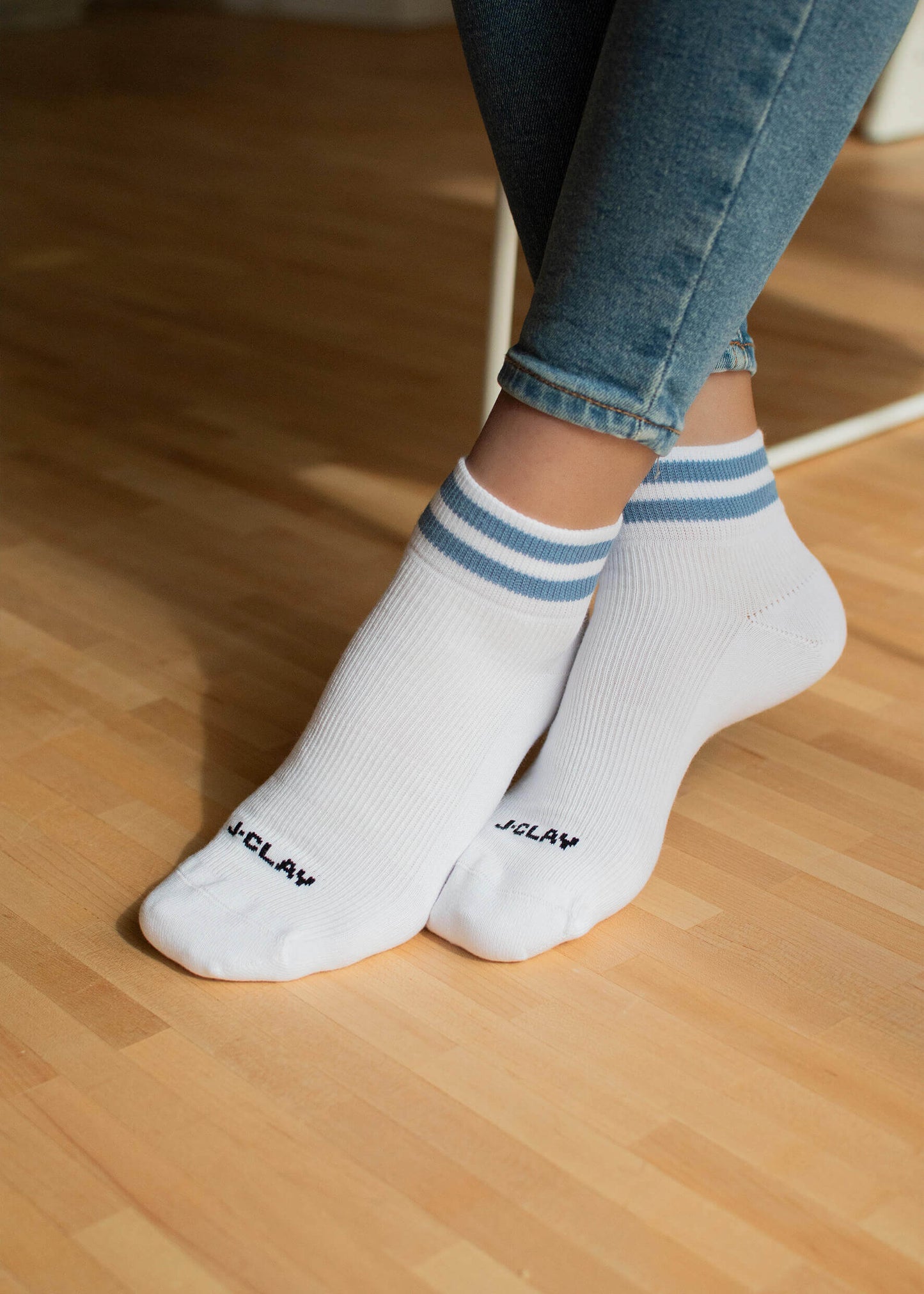 Eine Person in blauen Jeans sitzt mit gekreuzten Füßen auf einem hellen Holzboden und trägt J.Clay Sneaker Socken - weiße Söckchen mit blauen Streifen und kleiner schwarzer Schrift an den Zehen.