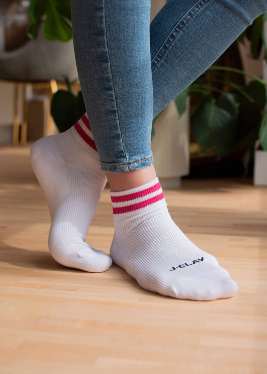Eine Person trägt J.Clay Sneaker Socken - weiß mit rosa Streifen und "J.Clay" in der Nähe der Zehen - mit blauen Jeans auf einem Holzboden. Im Hintergrund sind grüne Pflanzen und Möbel zu sehen.