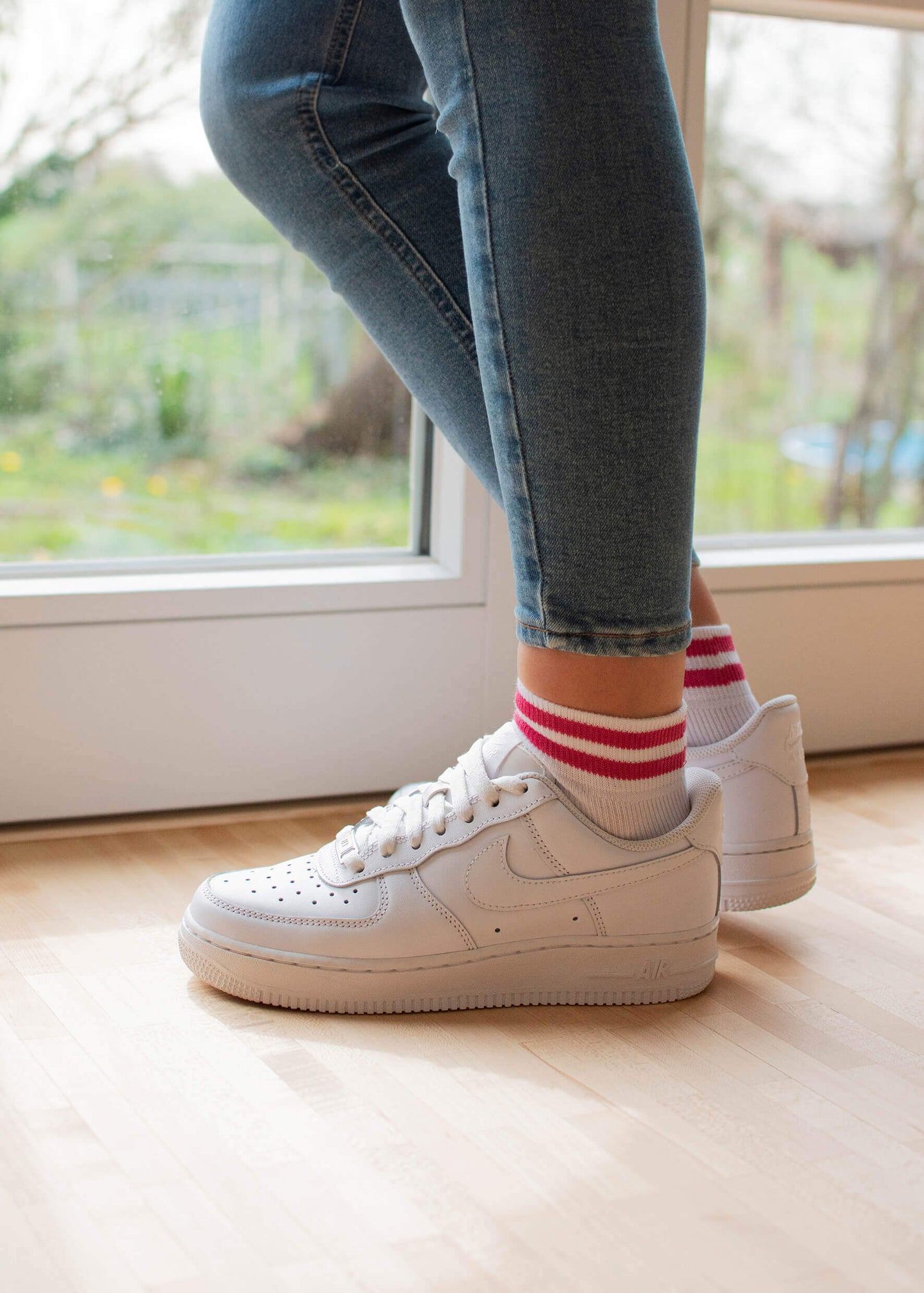Eine Person steht in einem Innenraum auf einem hellen Holzboden neben einer Glastür. Sie trägt eine blaue Skinny Jeans und weiße J.Clay Sneaker Socken mit roten Streifen.