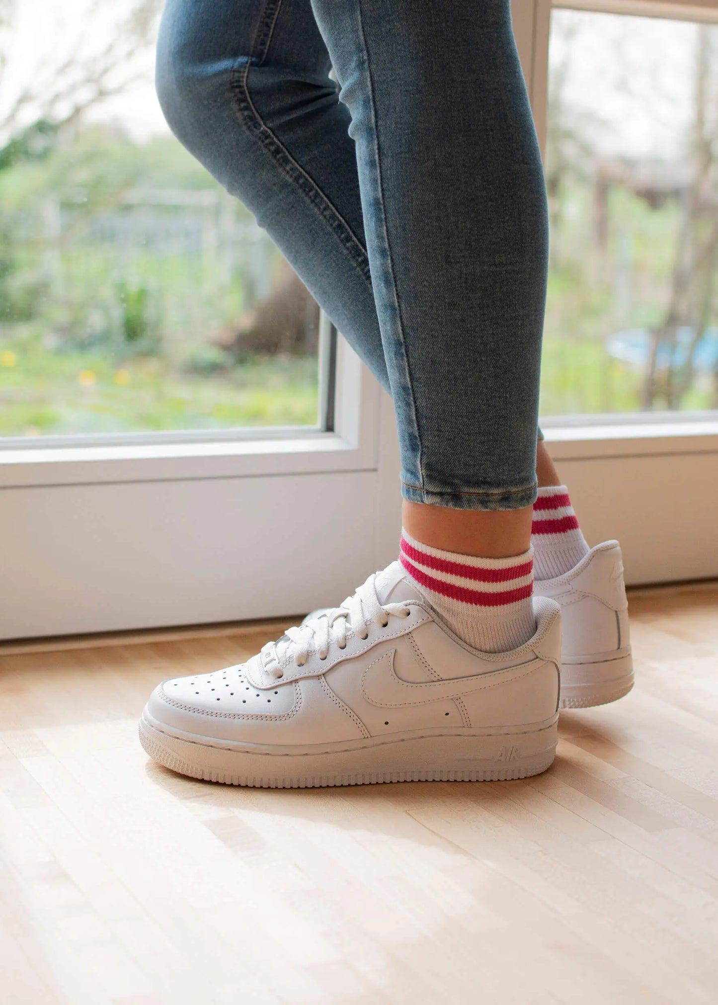 Eine Person steht in einem Innenraum auf einem hellen Holzboden neben einer Glastür. Sie trägt eine blaue Skinny Jeans und weiße J.Clay Sneaker Socken mit roten Streifen.