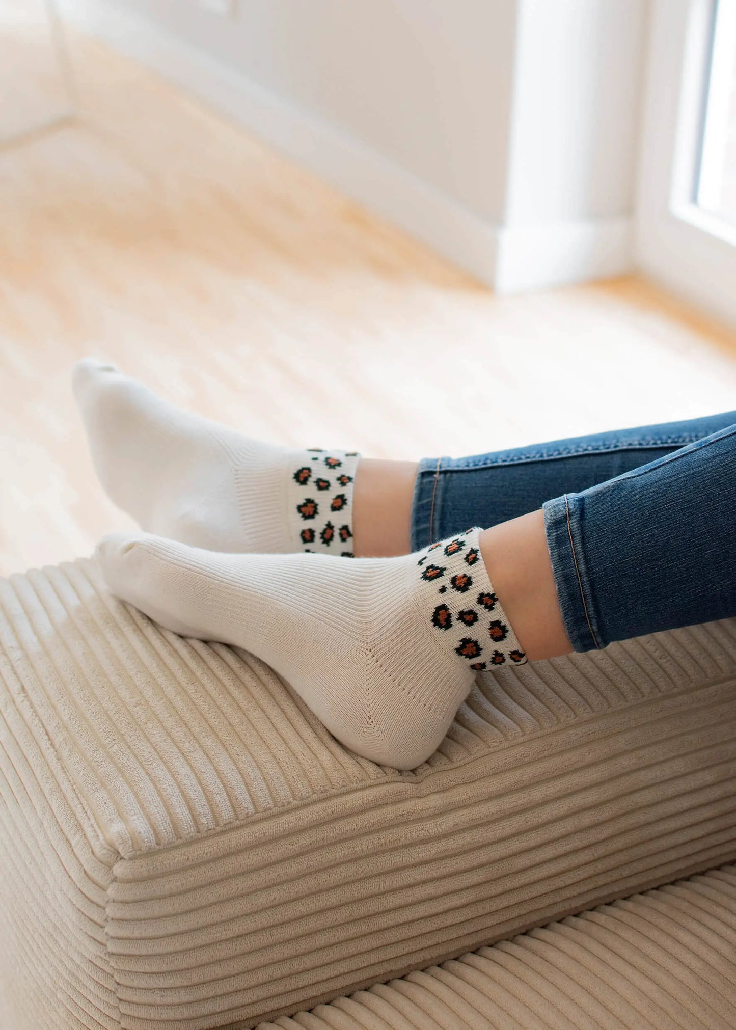 Eine Person in Sneaker-Socken mit Leopardenmuster von J.Clay mit weißen Bündchen und blauen Jeans ruht ihre Füße auf einem beigen, gerippten Ottomanen in einem hellen, modernen Raum.