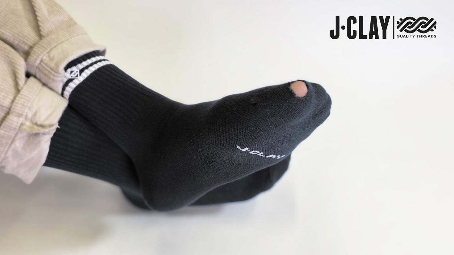 Eine Person trägt schwarze J-Clay-Socken mit einem sichtbaren Loch am großen Zeh. Das J Clay-Logo und der Text Quality Threads erscheinen in der oberen rechten Ecke auf weißem Hintergrund.