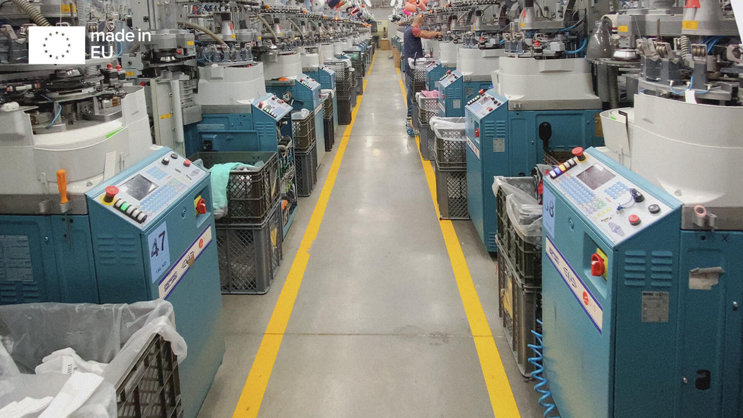 Reihen von blauen Industriestrickmaschinen säumen beide Seiten eines Fabrikgangs mit gelben Bodenmarkierungen. Jede Maschine hat Körbe und Schalttafeln. In der oberen linken Ecke ist ein "Made in EU"-Etikett angebracht.