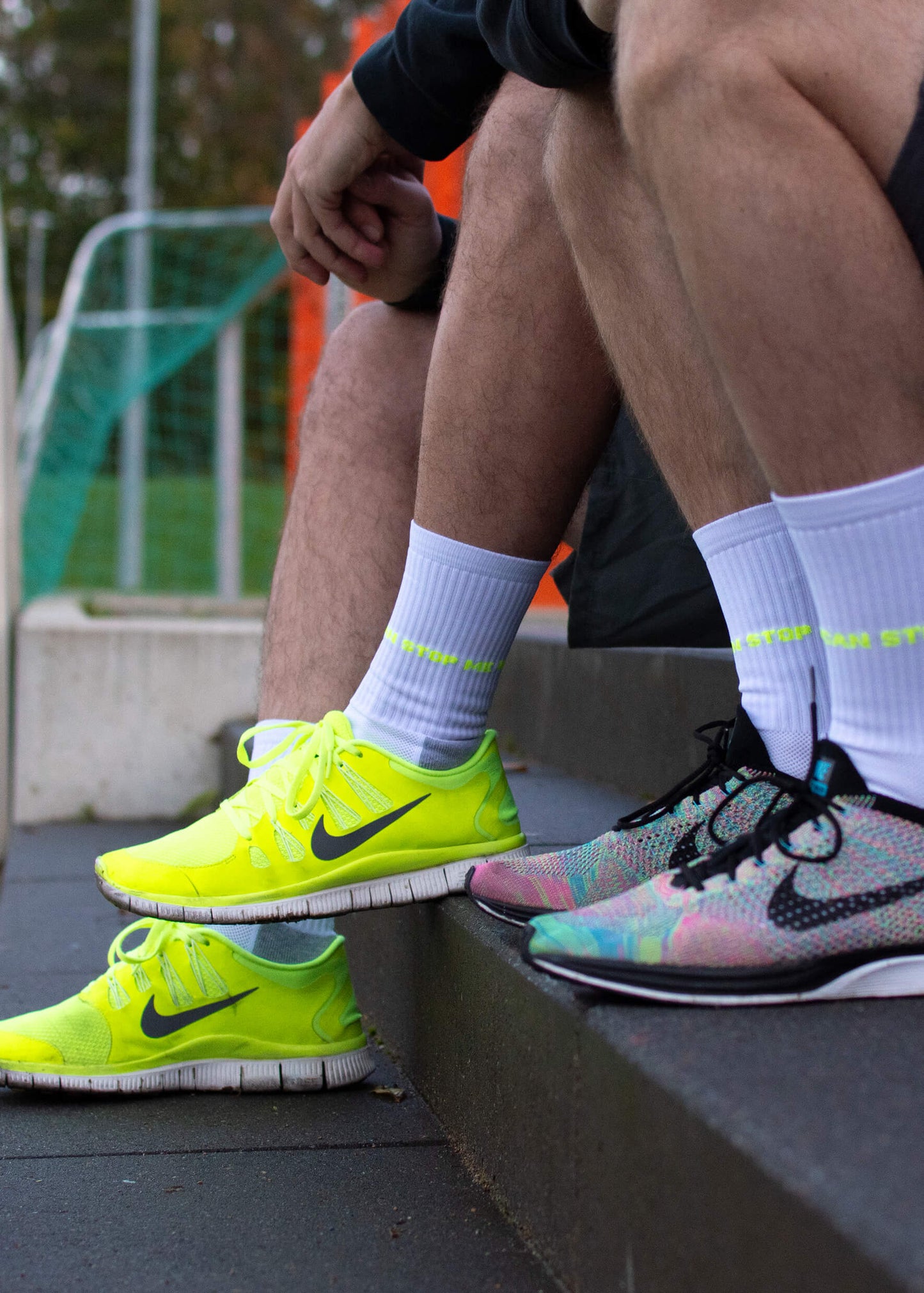 Zwei Personen sitzen auf Stufen im Freien und tragen weiße Sportsocken mit gepolsterter Sohle von J.Clay. Einer trägt leuchtend gelbe Turnschuhe, der andere bunte. Im Hintergrund ist ein Sportplatz zu sehen.