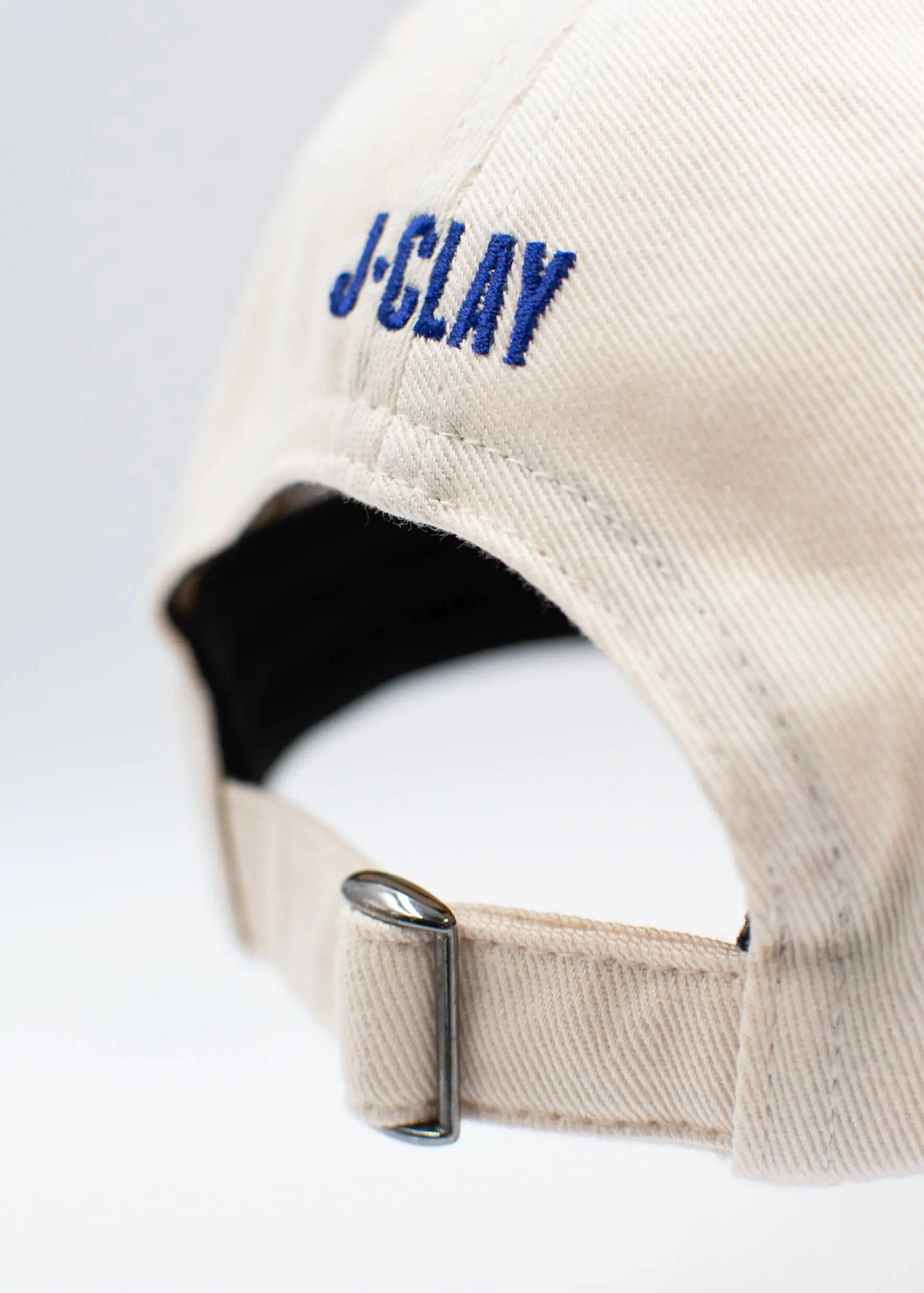Nahaufnahme der Rückseite einer beigen J.Clay Dad Cap mit blau gesticktem "J-CLAY" über dem verstellbaren Metallschnallenband.