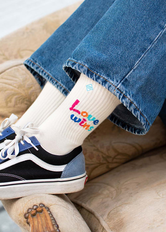 Person in Jeans, Turnschuhen und J.Clay „Love Wins“-Tennissocken mit bunten „Love Wins“-Buchstaben.
