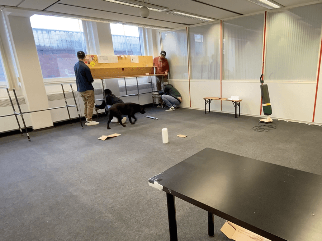 Vier Personen arbeiten in einem größtenteils leeren Büroraum zusammen und montieren oder arrangieren Gegenstände in der Nähe der Fenster. Ein schwarzer Hund läuft in der Nähe herum, und auf dem Teppichboden liegen verstreute Pappstücke und Reinigungsmittel.