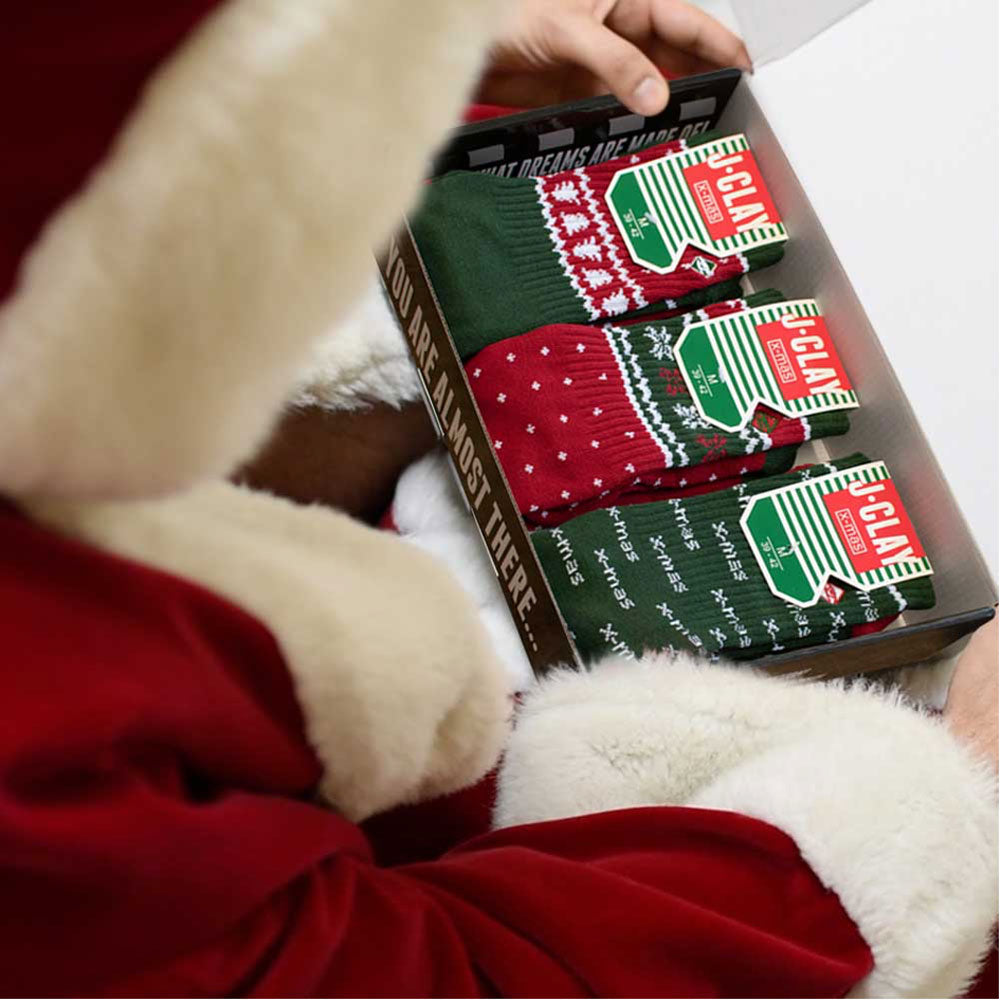 Eine Person in einem rot-weißen Weihnachtsmannkostüm hält eine Schachtel mit festlichen Socken mit grünen und roten Motiven und weihnachtlichen Mustern.