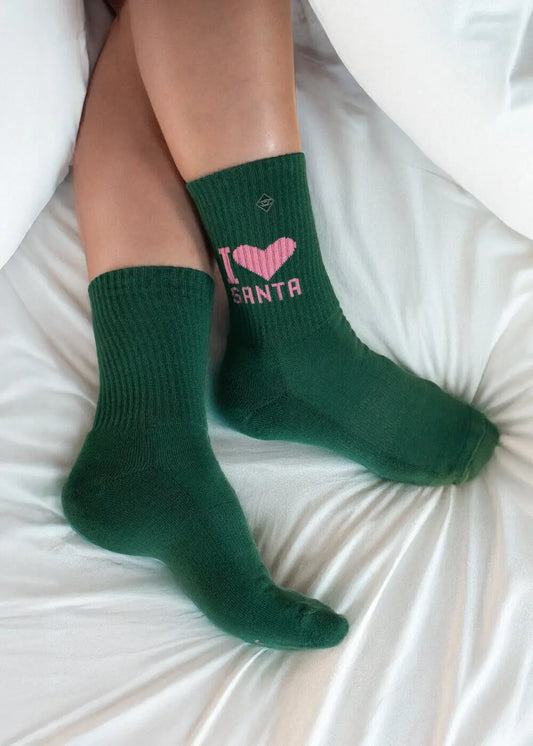 Sie liegt auf einem weißen Bettlaken und trägt grüne „J.Clay I Love Santa – Fam Pack“-Socken.