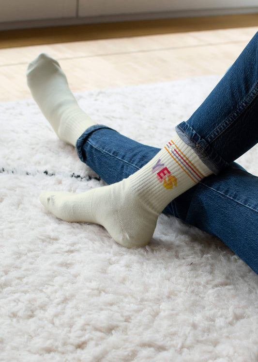 Eine Person sitzt im Schneidersitz auf einem weißen, flauschigen Teppich, trägt Blue Jeans und helle Tennissocken von J.Clay mit Regenbogenstreifen und dem Wort YES in bunten Buchstaben.