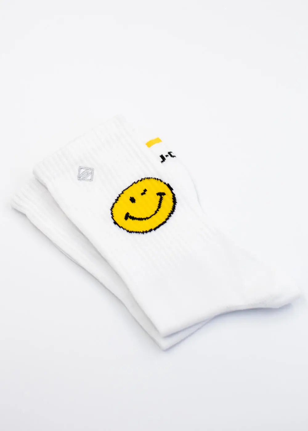 Ein Paar weiße Tennissocken von J.Clay Yellow Smile, entworfen in Deutschland, mit einer großen gelben Smiley-Grafik an der Seite und ordentlich gefaltet auf weißem Hintergrund.
