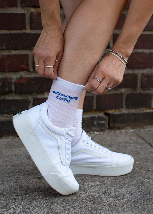 Jemand, der J.Clay trägt, ist immer zu spät – Midi-Socken und weiße Sneaker, hockt an einer Backsteinmauer.