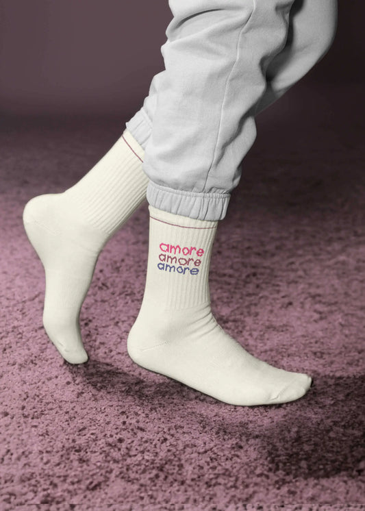 Eine Person in hellgrauer Jogginghose steht auf Zehenspitzen auf einem lilafarbenen Strukturteppich und trägt weiße J.Clay-Tennissocken, auf denen das Wort "amore" dreimal in Rosa und Lila aufgedruckt ist.
