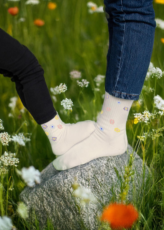 Zwei Personen stehen auf einem Felsen inmitten von Wildblumen und tragen weiße J.Clay-Socken mit bunten Blumenmustern. Ihre Füße berühren sich - einer trägt eine schwarze Hose, der andere eine blaue Jeans. Das Design ist inspiriert von J.Clay Baby Socken und Kinder Socken 0-4 Jahre.