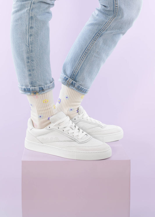 Eine Person steht auf einem pastellrosa Podest vor einem hellvioletten Hintergrund. Sie trägt eine hellgewaschene, hochgekrempelte Jeans, cremefarbene Tennissocken von J.Clay mit kleinen bunten Mustern und weiße Turnschuhe.