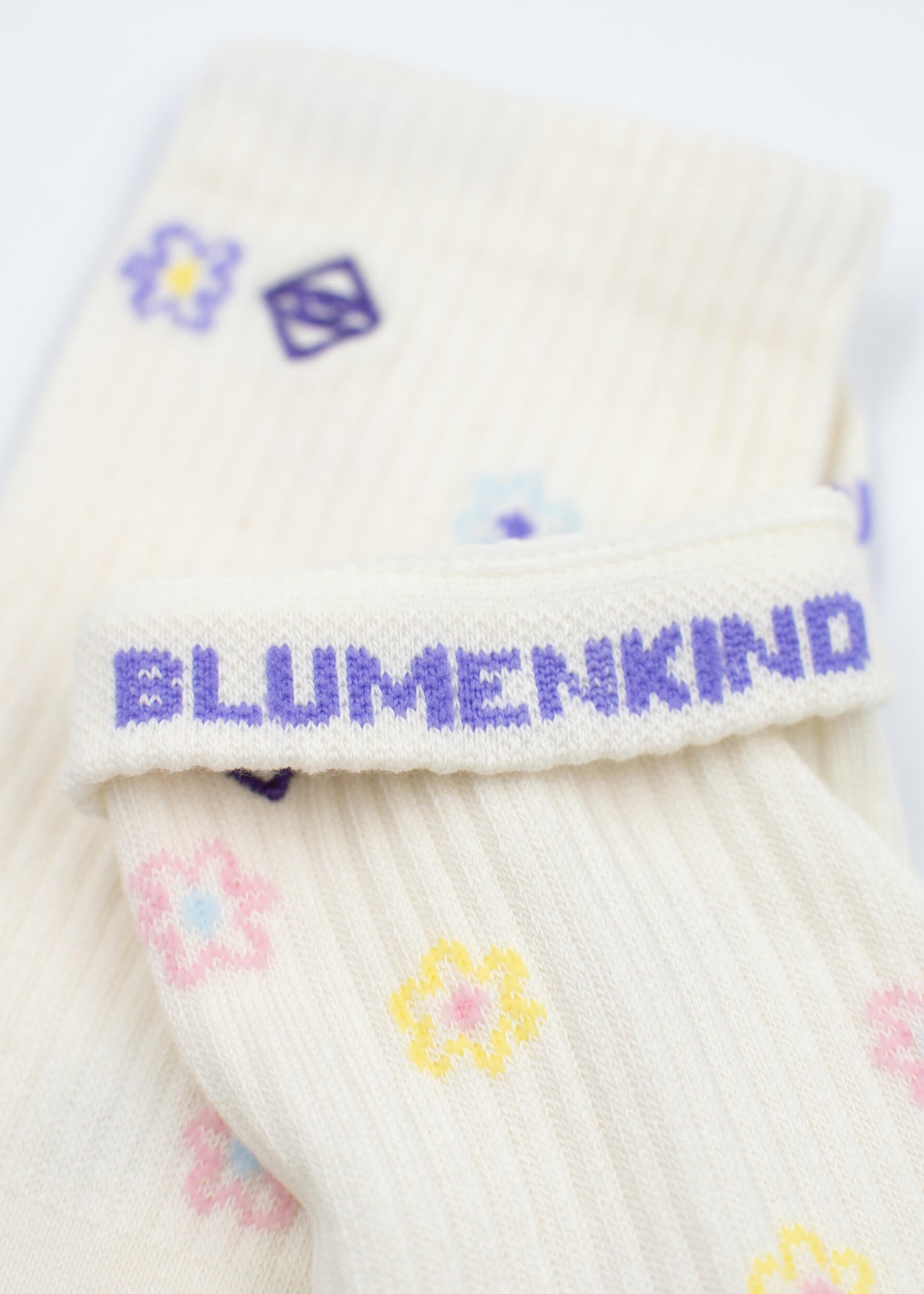 Eine Nahaufnahme der cremefarbenen Tennissocken von J.Clay, die mit pastellfarbenen Blumenmustern verziert sind und auf dem Bündchen den Schriftzug BLUMENKIND" in Lila tragen.