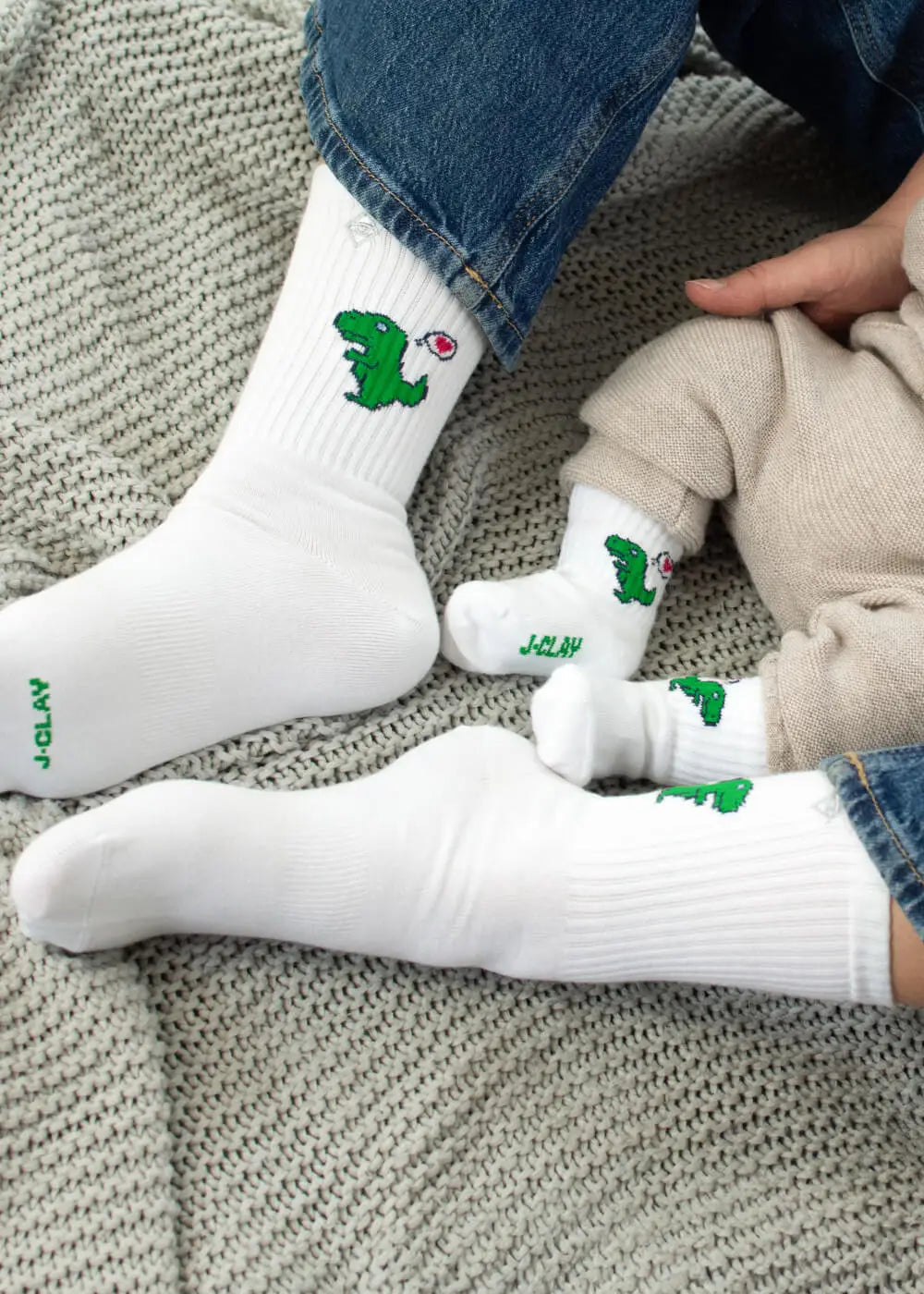 Ein Erwachsener und ein Baby sitzen auf einer grauen Strickdecke, beide tragen passende J.Clay Mama Kind Socken - weiße Socken mit grünen Cartoon-Dinosauriern. Der Erwachsene kombiniert sie mit einer blauen Jeans, das Baby trägt eine beigefarbene, so dass ein perfekter Partnerlook entsteht.