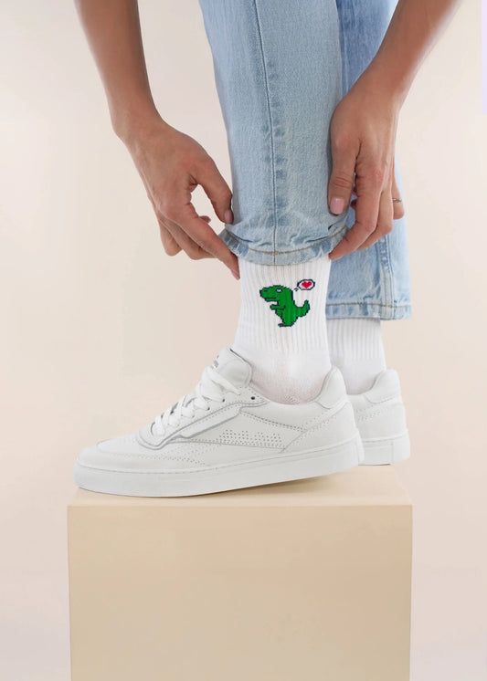 Eine Person in hellblauen Jeans und weißen Turnschuhen krempelt ihr Hosenbein hoch und zeigt die trendigen J.Clay Tennissocken mit einem grünen Dinosaurier und einem roten Herz, die auf einer beigen Schachtel stehen.