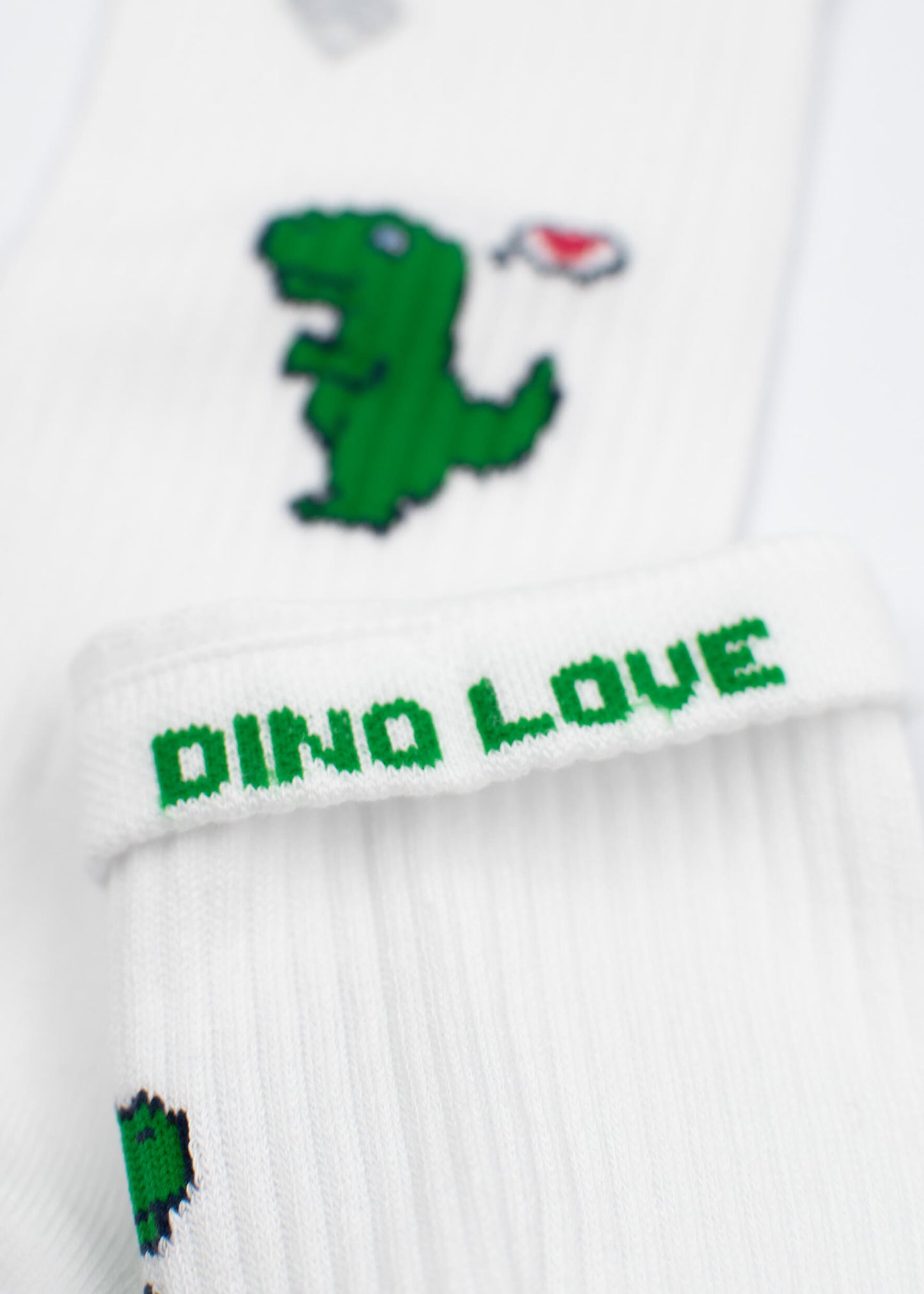 Nahaufnahme einer weißen gerippten Tennissocke von J.Clay mit einem grünen Cartoon-Dinosaurier, einem Herz in der Nähe seines Mundes und "DINO LOVE" in grüner Farbe auf dem Bündchen.