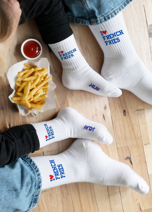 Zwei Menschen in Jeans sitzen eng aneinander gedrängt auf einem Holzboden und tragen weiße J.Clay I ❤️ French Fries Baby Socken. Pommes Frites und Ketchup liegen daneben - niedliche Kinder Socken für 0-4 Jahre, perfekt für die Kleinsten.