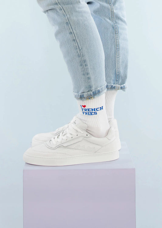Eine Person in hellblauen Jeans mit Bündchen, weißen Turnschuhen und J.Clay-Tennissocken mit dem Schriftzug "I ❤️ FRENCH FRIES" steht auf einem pastellfarbenen lila Würfel vor hellblauem Hintergrund.