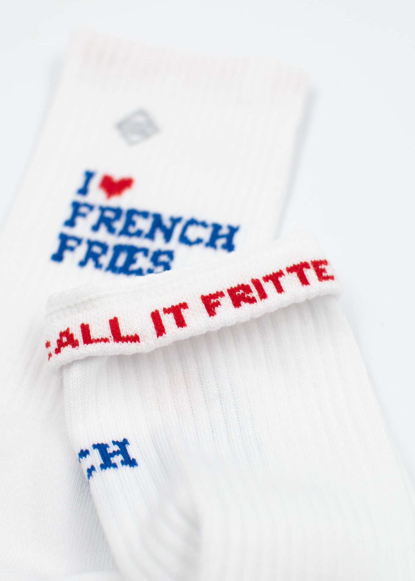 Eine Nahaufnahme von weißen J.Clay Tennissocken mit blauem Text "I ♥ FRENCH FRIES" und rotem Manschettentext "CALL IT FRITTE", mit einem gerippten Design, dargestellt auf einer weißen Fläche.
