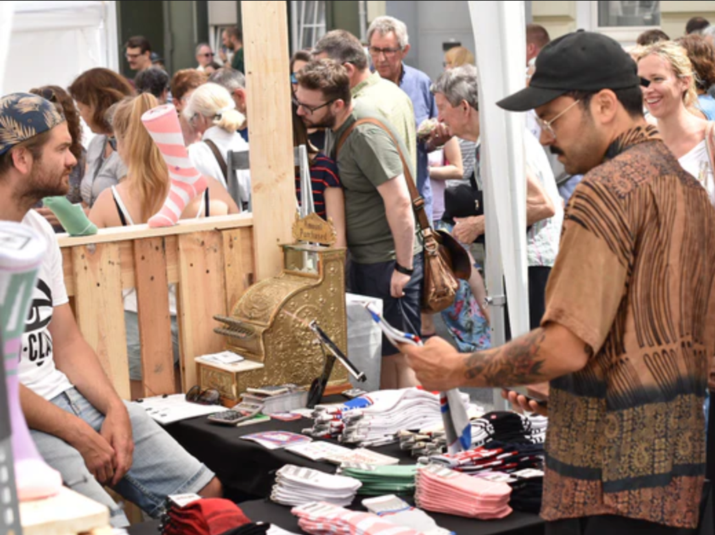 Eine belebte Marktszene im Freien mit Menschen, die an einem Verkaufsstand nach Socken suchen und sie kaufen. Ein Mann sitzt hinter einem Tisch, während ein anderer Socken begutachtet, umgeben von Waren und einer altmodischen Kasse.