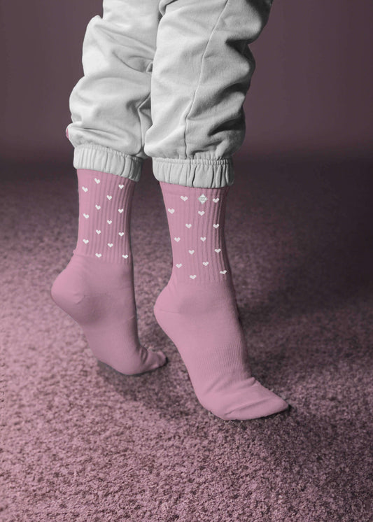 Eine Person steht auf Zehenspitzen in hellrosa Tennissocken von J.Clay mit kleinen weißen Herzen und hellgrauen Hosen mit Bündchen auf einem weichen rosa Teppich vor einem rosa Hintergrund.