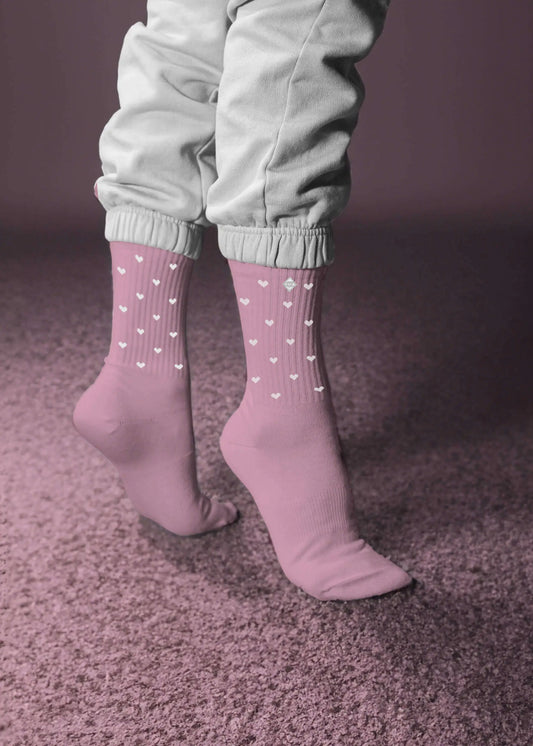 Eine Person steht auf Zehenspitzen in hellrosa Tennissocken von J.Clay mit kleinen weißen Herzen und hellgrauen Hosen mit Bündchen auf einem weichen rosa Teppich vor einem rosa Hintergrund.